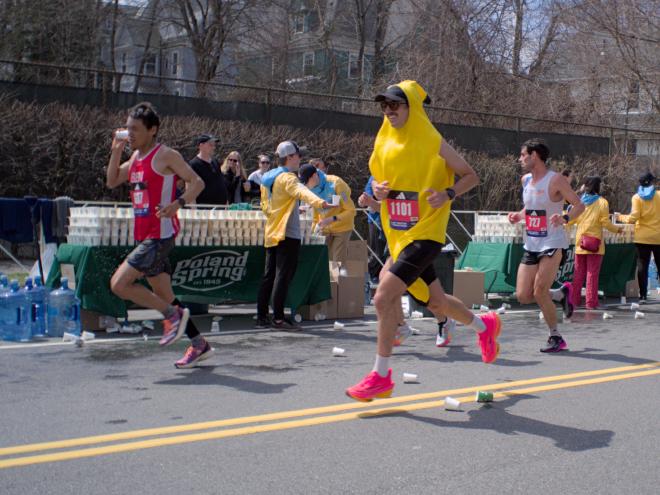 A man dressed in a banana costume with pink shoes runs past a water station.