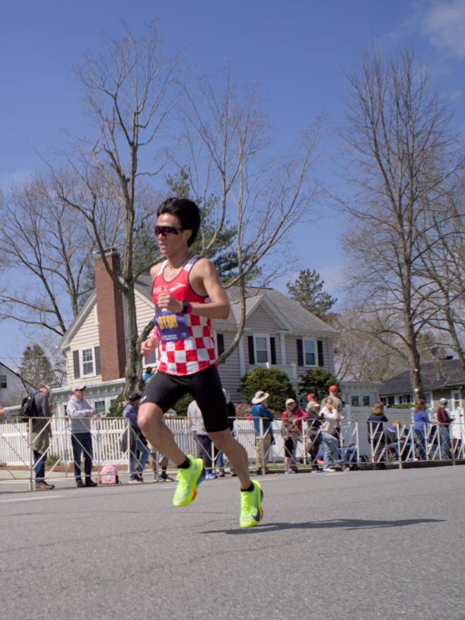 A man wearing a red and white checkered top and neon green running shoes. His short dark hair is bouncing with his stride.